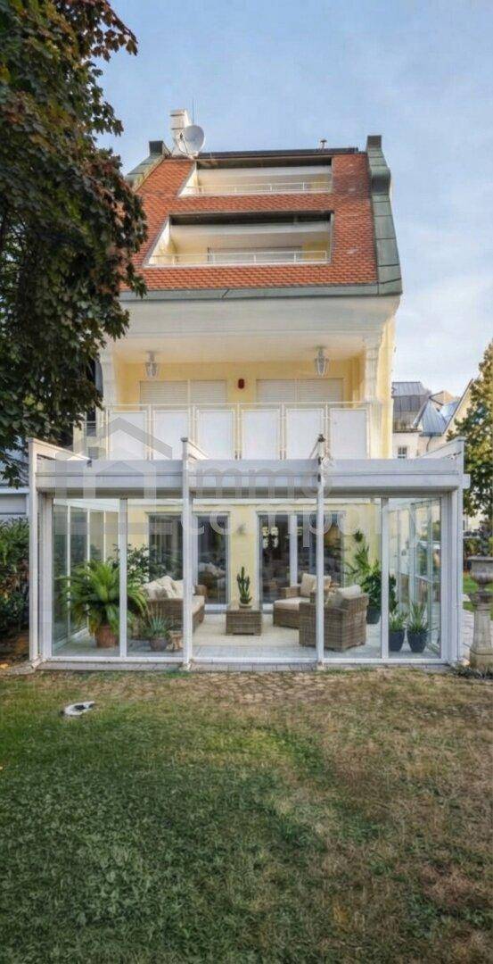 Rückansicht eines Hauses mit einem modernen Wintergarten und mehreren Balkonen, umgeben von einem Rasen.