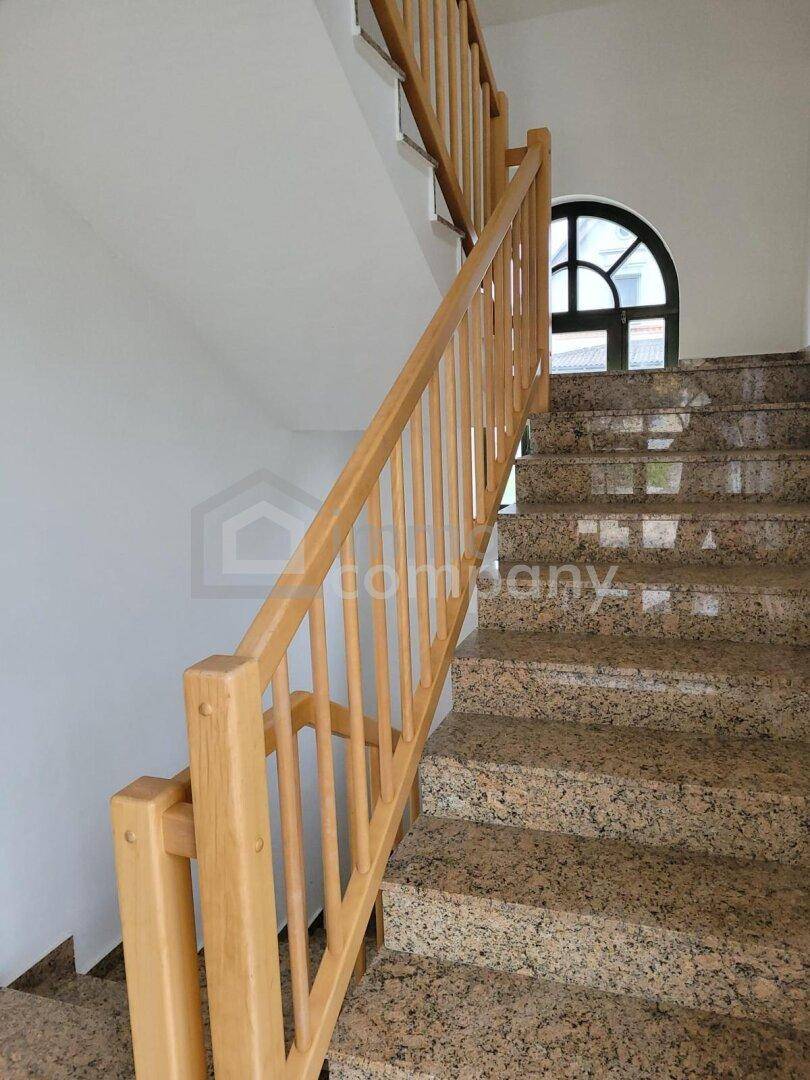 Heller Treppenaufgang mit Holzgeländer und Granitstufen, führt in die oberen Stockwerke.