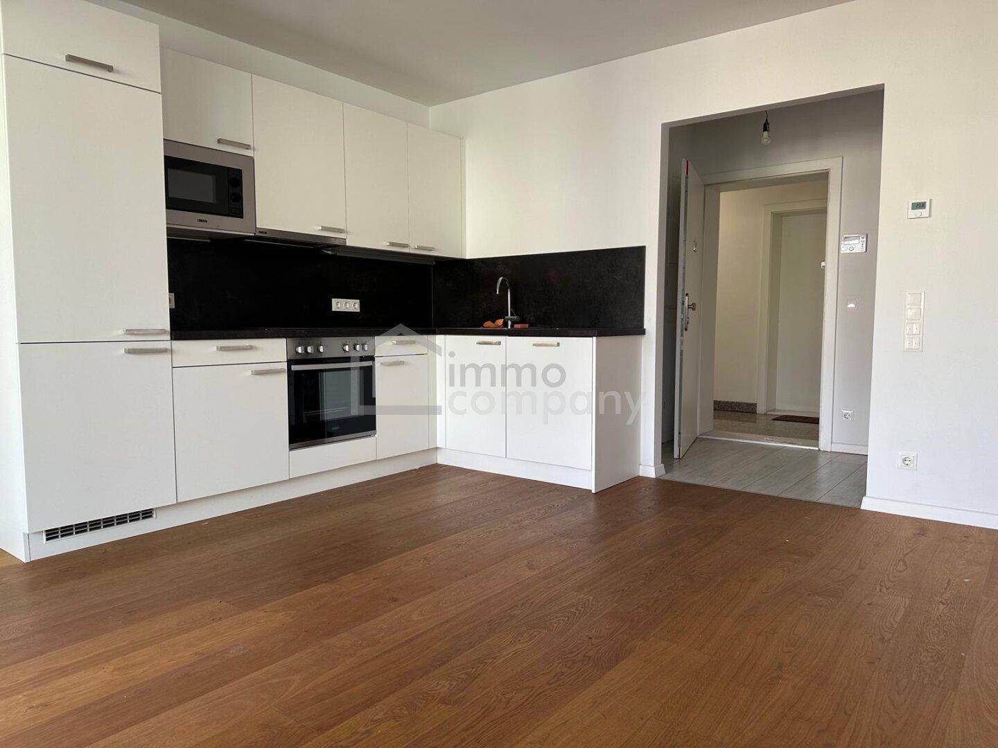 Heller, leerer Wohnbereich mit integrierter weißer Küche und hochwertigem Holzboden.