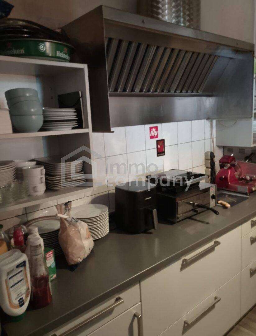 Professionelle Gastroküche mit Edelstahl-Dunstabzugshaube, Arbeitsflächen und Regalen für Geschirr.