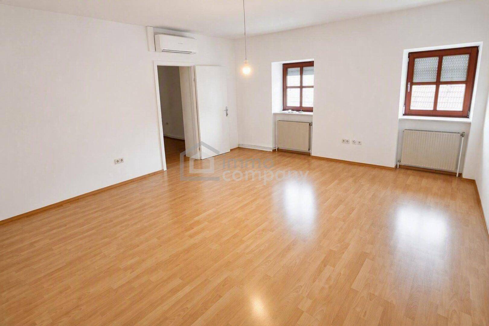 Lichtdurchflutetes Zimmer mit Holzboden, zwei Fenstern und Zugang zu einem weiteren Raum.