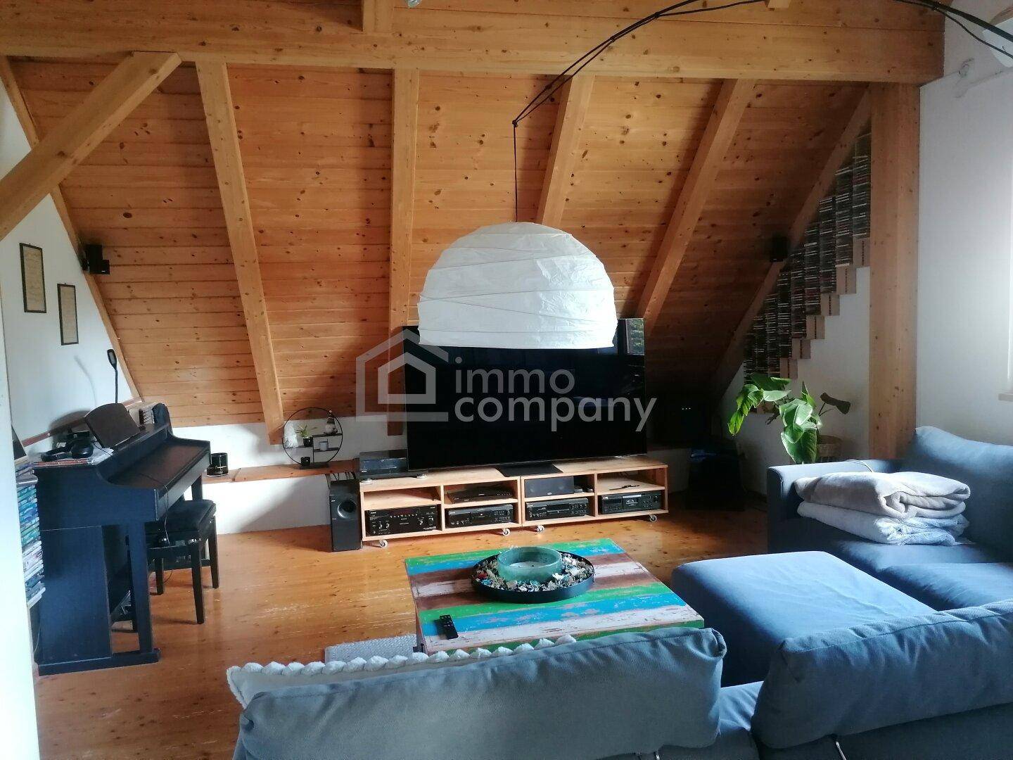 Gemütliches Wohnzimmer mit Holzbalkendecke, großem Sofa und einem Klavier unter der Dachschräge.