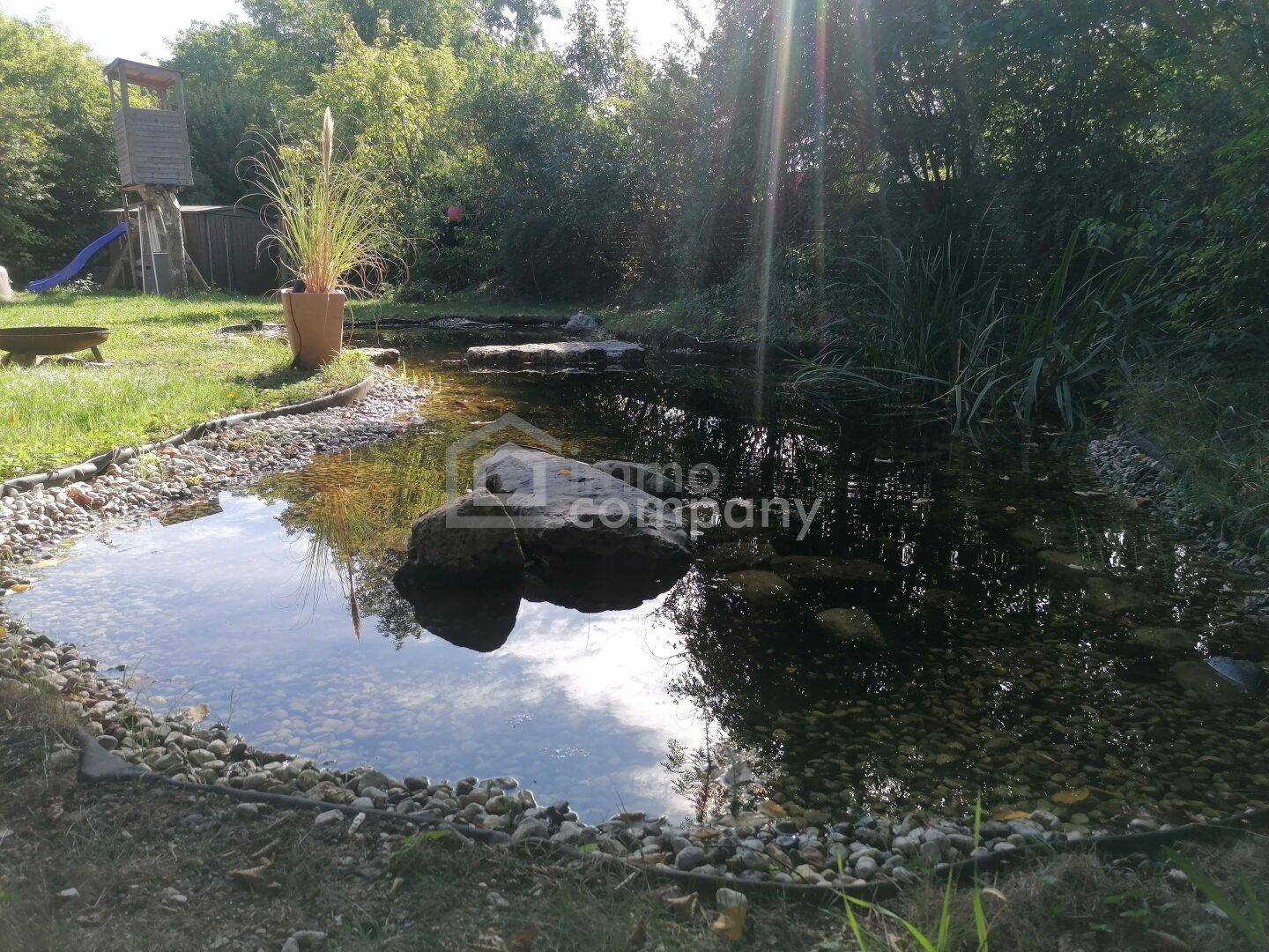 Sonniger Gartenbereich mit einem natürlichen Teich, umgeben von Bäumen und einem Spielturm.