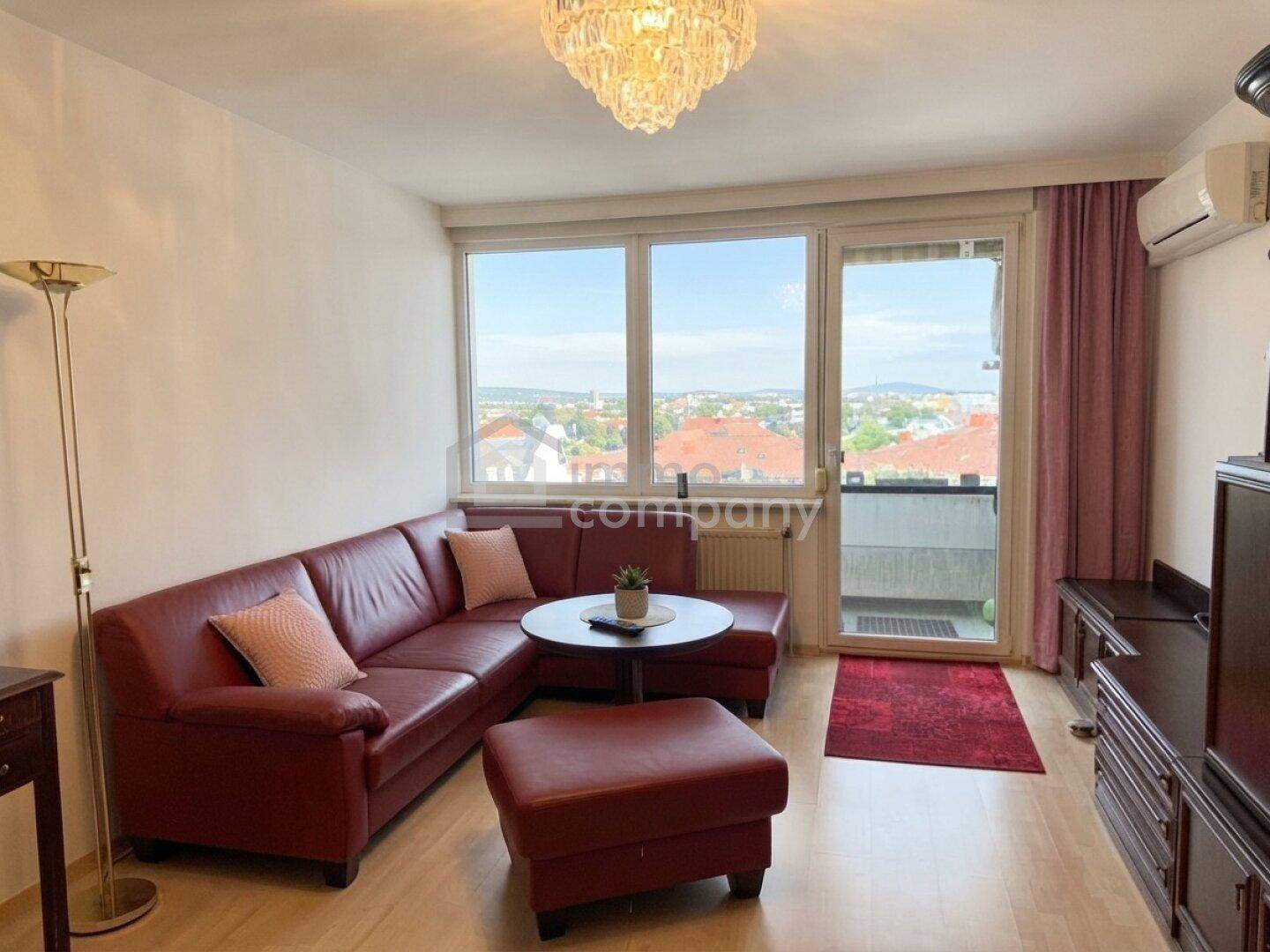Helles Wohnzimmer mit rotem Ledersofa, Klimaanlage und Zugang zum Balkon mit Stadtblick.