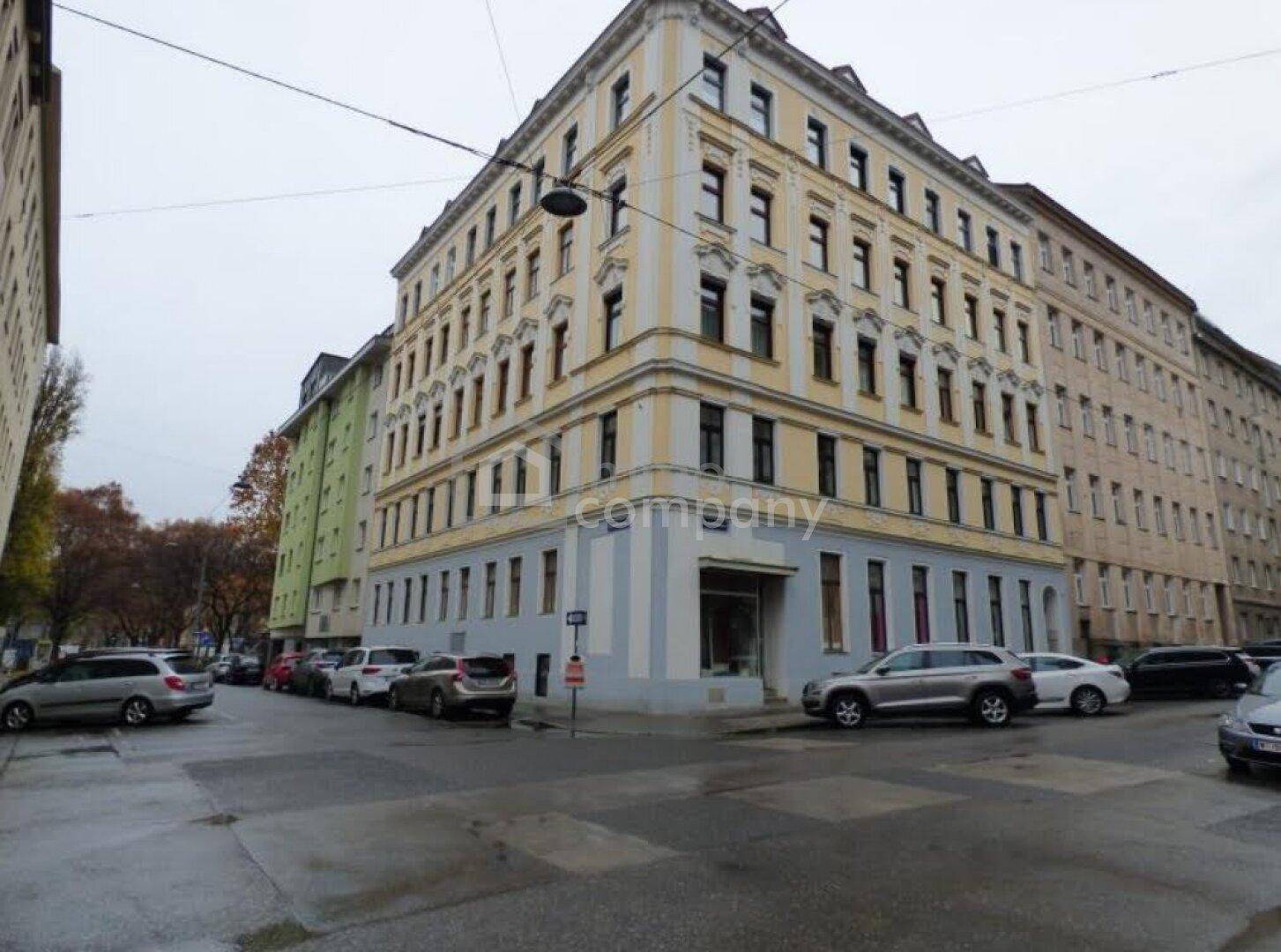 Historisches Eckgebäude mit gelb-grauer Fassade an einer belebten Stadtstraße mit parkenden Autos.