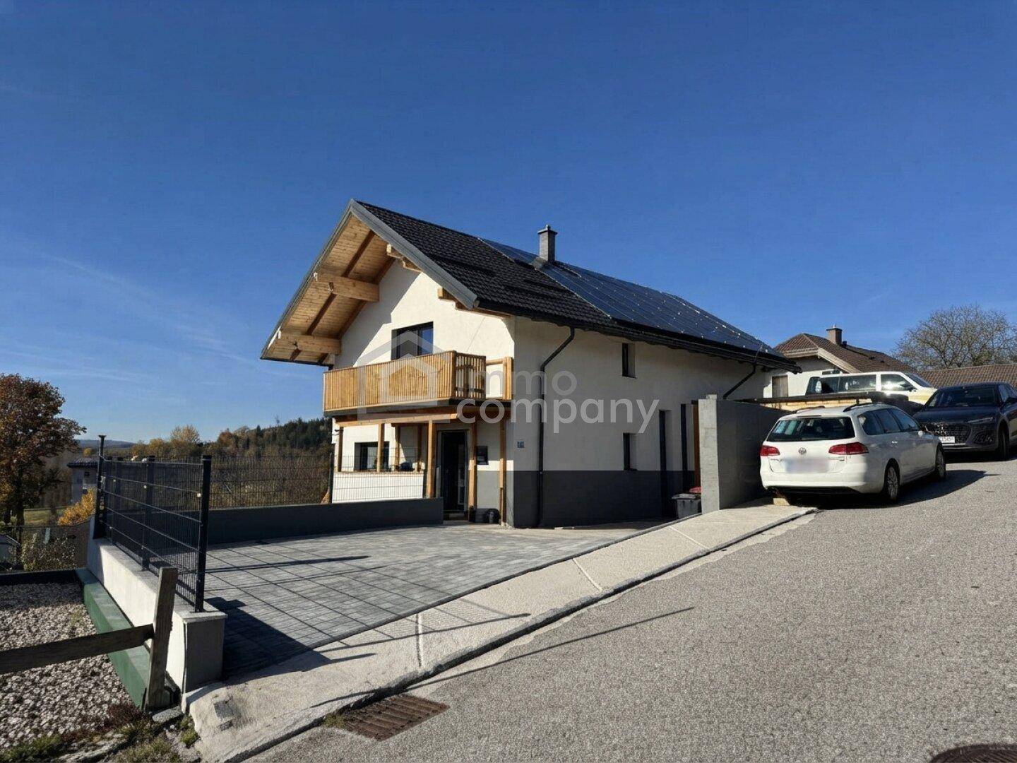 Modernes Einfamilienhaus mit Photovoltaikanlage, Holzbalkon und gepflasterter Auffahrt, umgeben von grüner Landschaft.