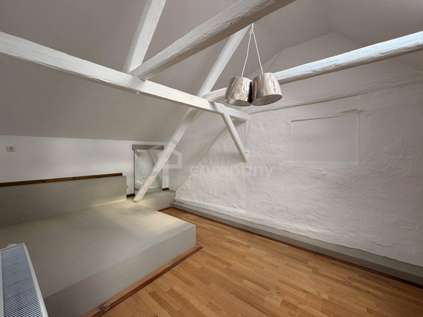 Helles Schlafzimmer im Dachgeschoss mit sichtbaren Holzbalken, schrägen Wänden und Holzboden.
