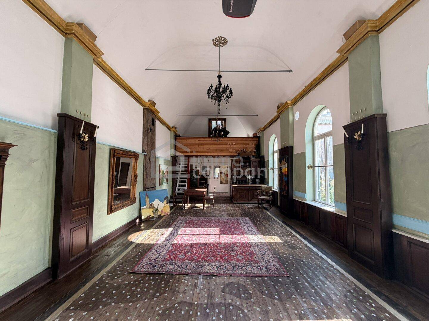 Großer, hoher Raum mit Holzboden, Stuckelementen und einem Kronleuchter, der als Galerie genutzt wird.