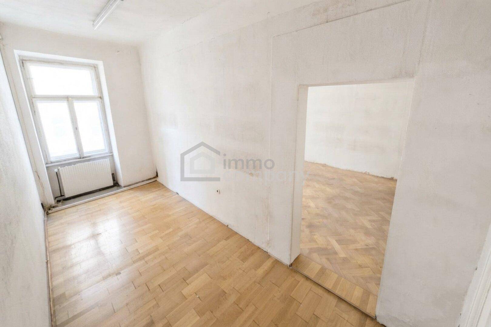 Heller, leerer Raum mit Parkettboden und einem großen Fenster, offen zu einem Nebenraum.