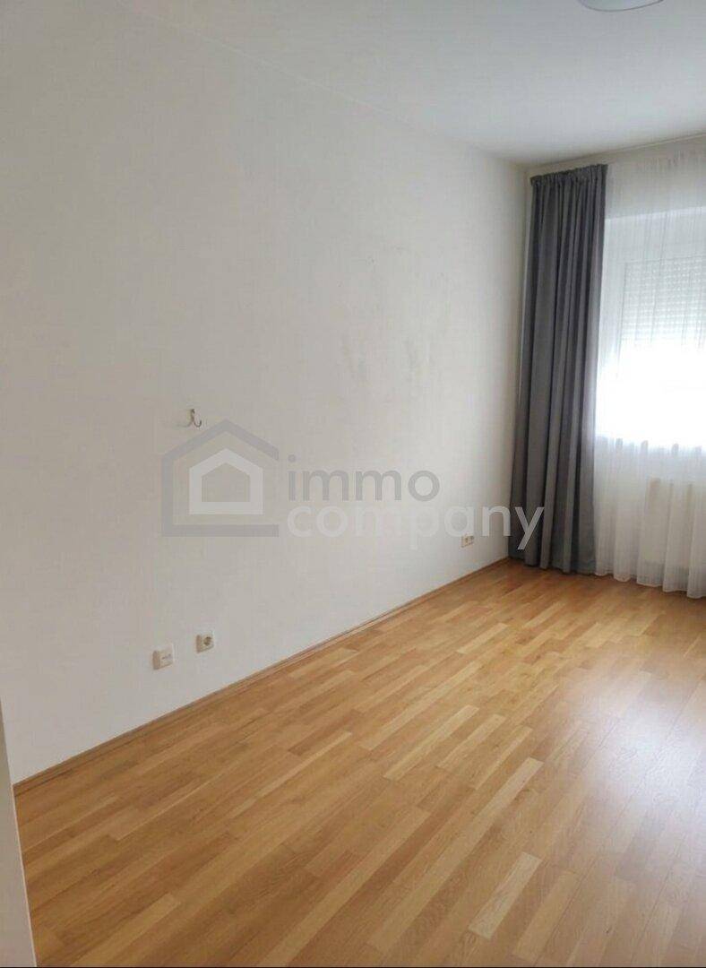 Leeres Zimmer mit hellem Holzboden, weißen Wänden und einem Fenster mit grauen Vorhängen, bereit zur Einrichtung.