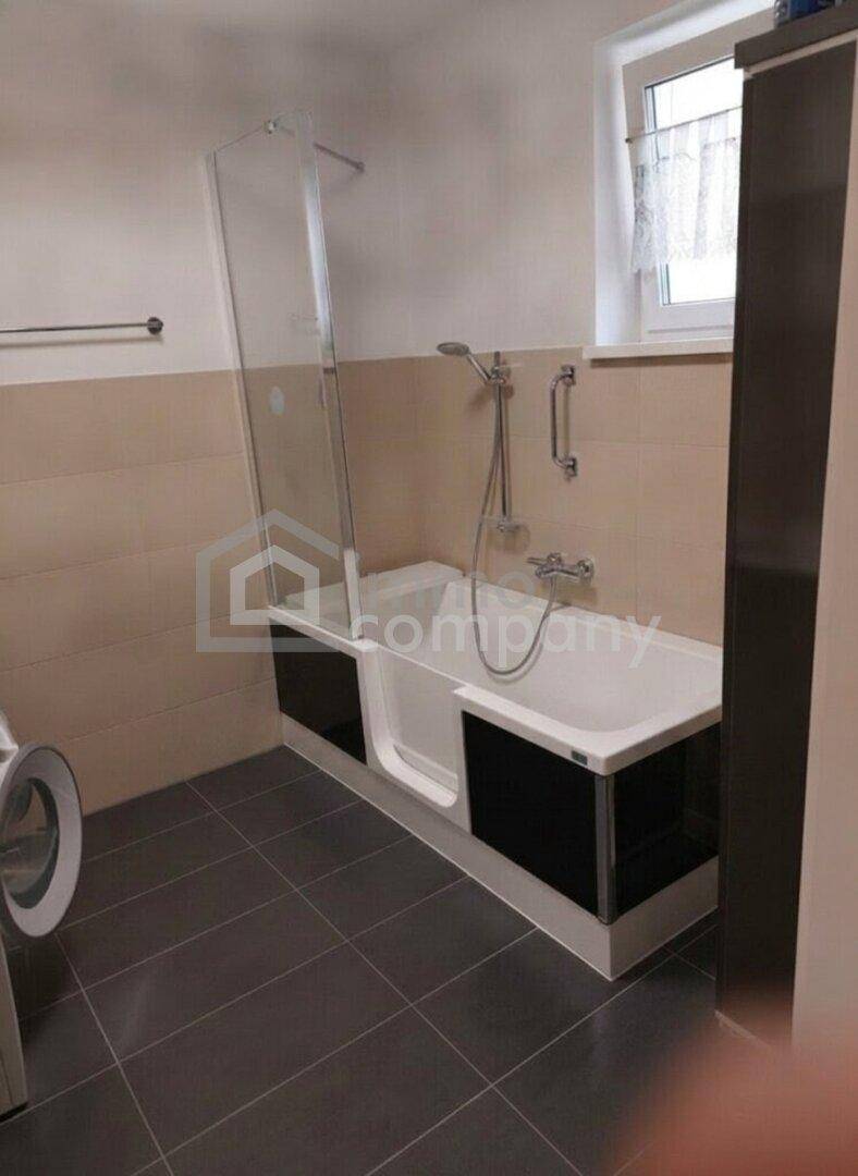 Badezimmer mit Badewanne inklusive Duschabtrennung und Fenster, hell gefliest und praktisch gestaltet.