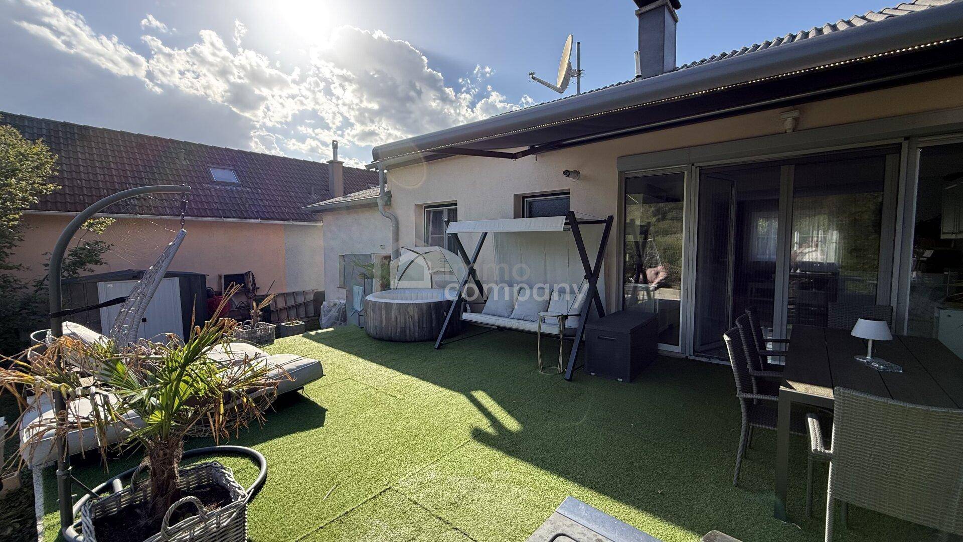 Geräumige Terrasse mit Kunstrasen, Whirlpool, Schaukel und Essbereich unter blauem Himmel.