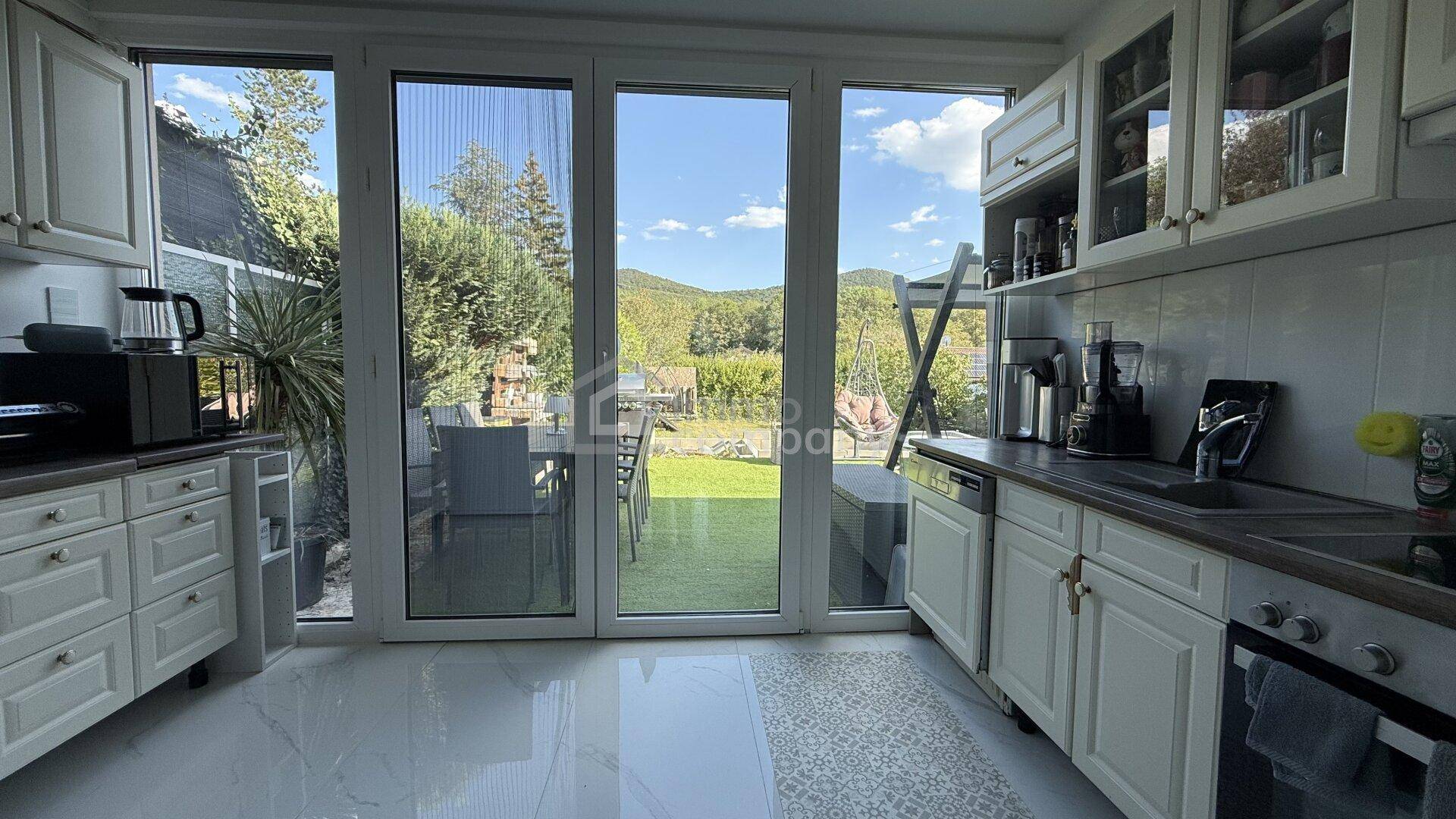 Helle Küche mit weißen Schränken, Fliesenboden und großem Fenster mit Blick ins Grüne.