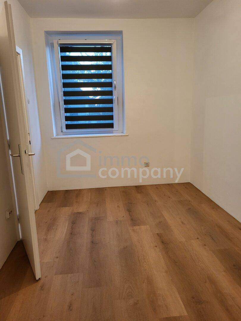 Unmöbliertes Zimmer mit Laminatboden und einem Fenster mit horizontalen Jalousien.