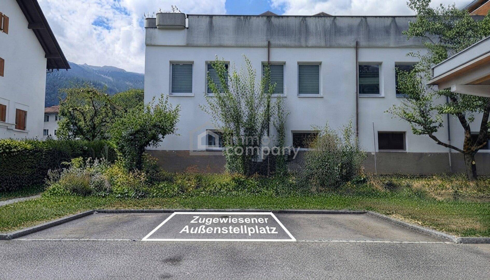 Zugewiesener Außenstellplatz vor einem Wohngebäude mit grüner Umgebung und Bergblick unter blauem Himmel.