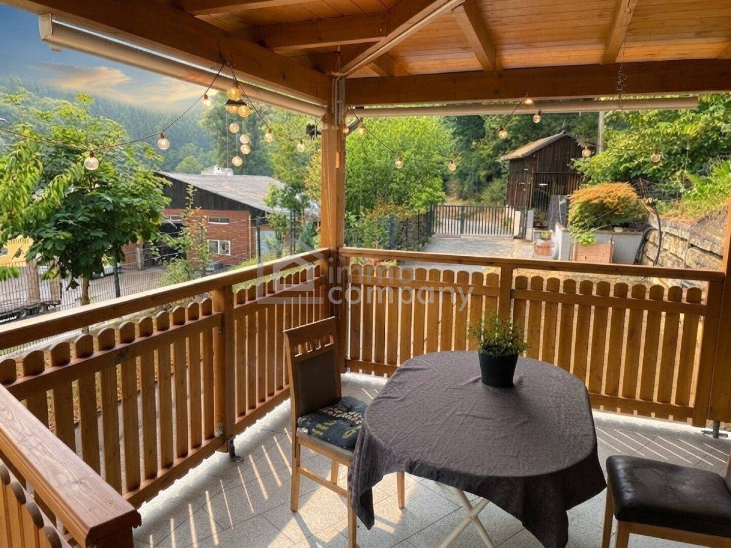 Gemütliche überdachte Terrasse mit Sitzgelegenheiten und Lichterkette, umgeben von grüner Landschaft.