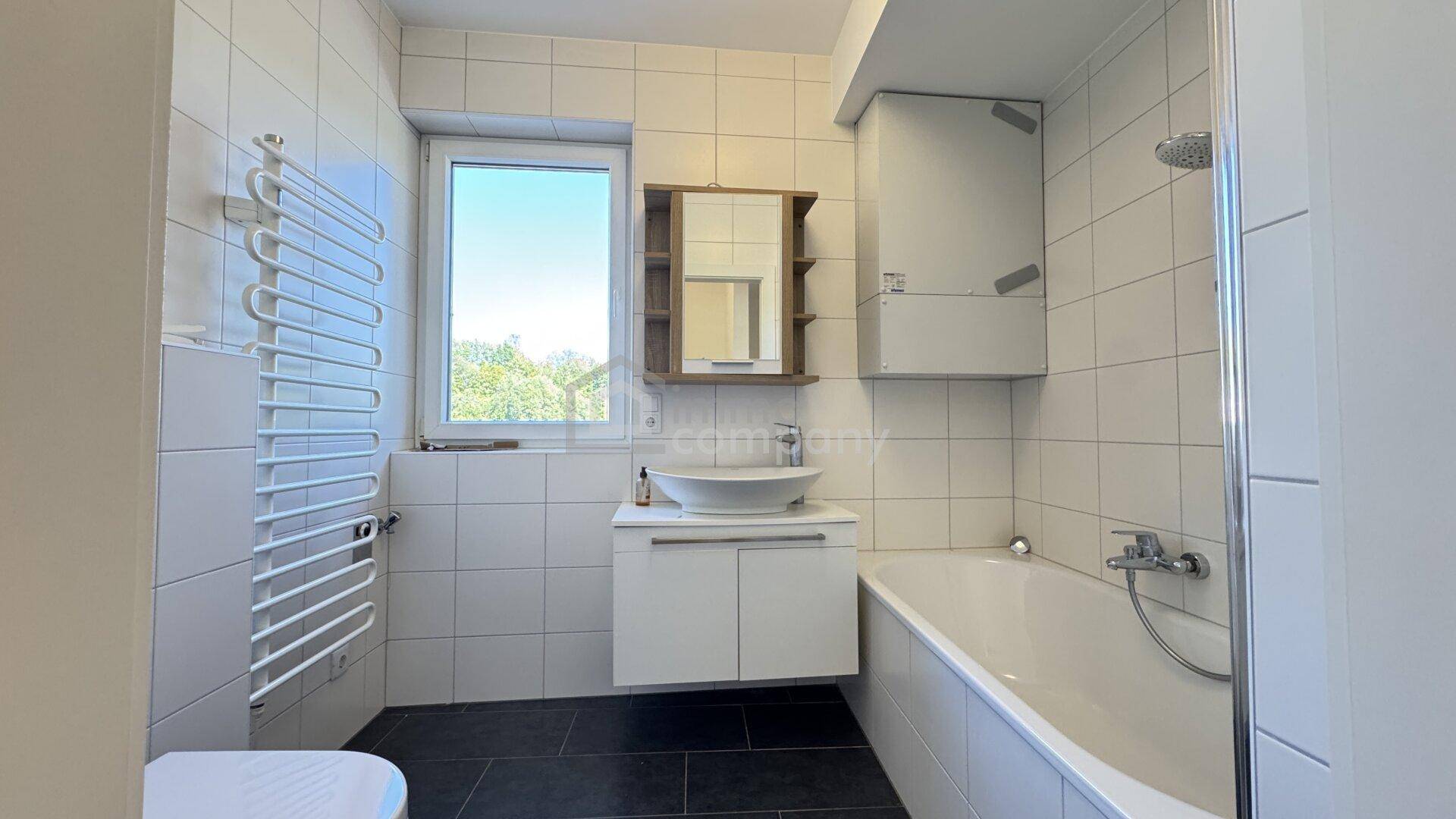 Modernes Badezimmer mit weißen Wandfliesen, dunklem Boden und Badewanne sowie Fenster.