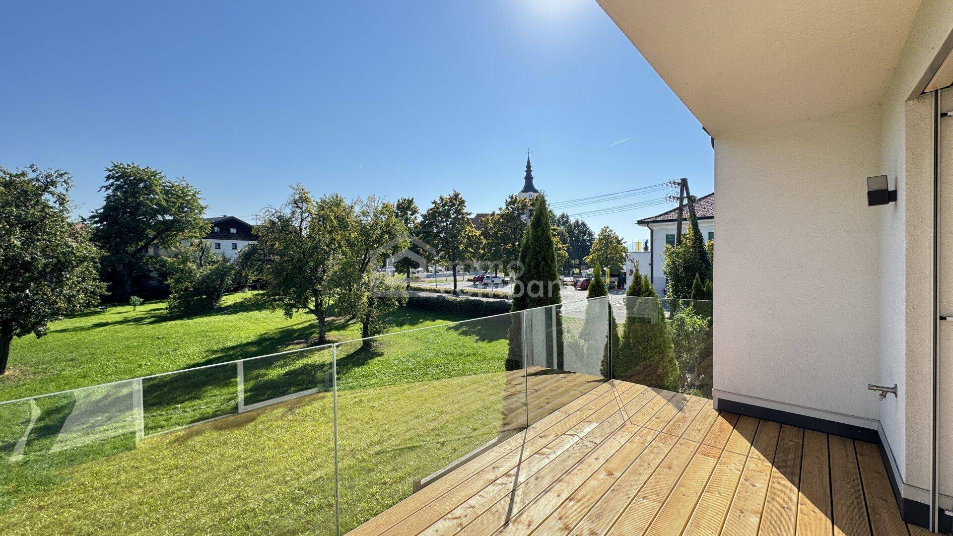 Sonniger Balkon mit Holzboden und Glasbrüstung, mit Blick auf eine Grünfläche und einen Kirchturm.
