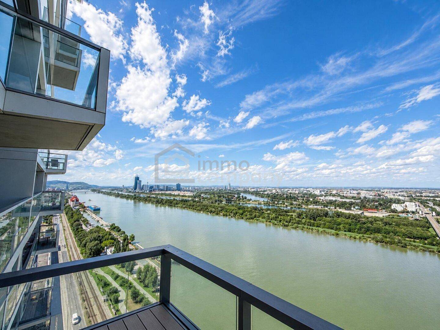 Weitläufiger Balkonblick auf den Fluss, die Stadt und die Berge unter strahlend blauem Himmel.
