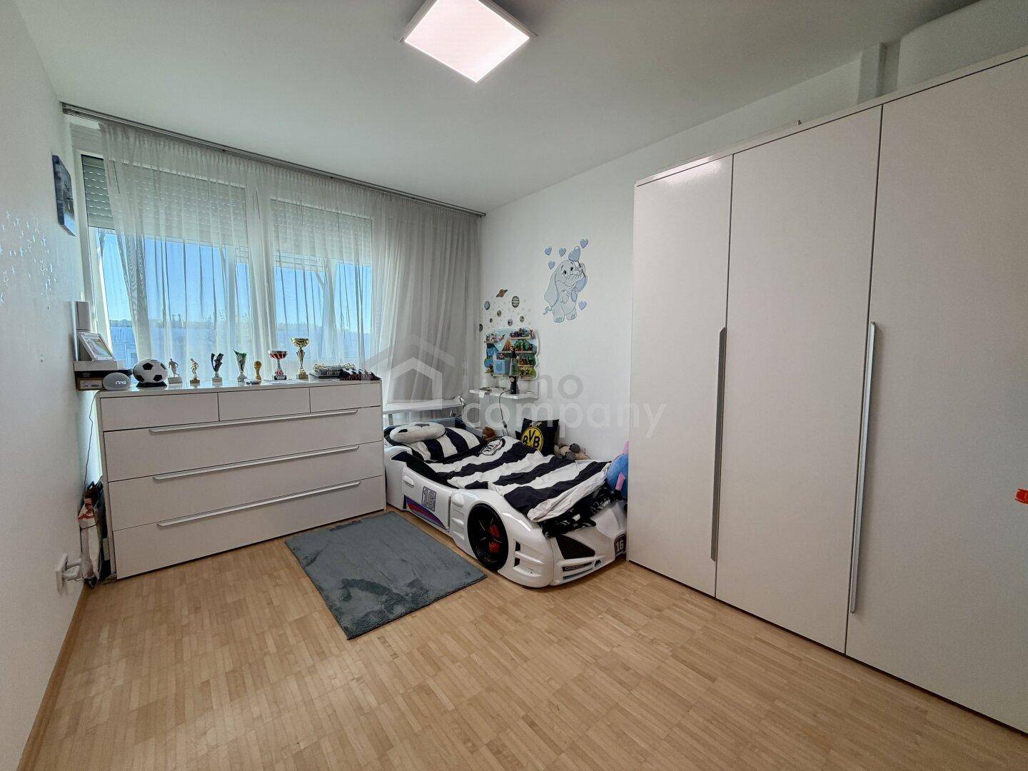 Kinderzimmer mit hellem Holzboden, großem Fenster und weißem Einbauschrank.