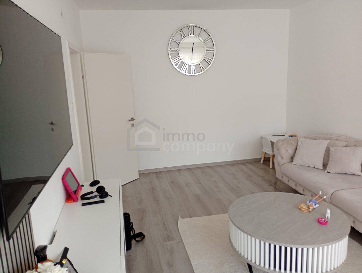 Geräumiges Wohnzimmer mit großem Fernseher, Wanduhr und Laminatboden.
