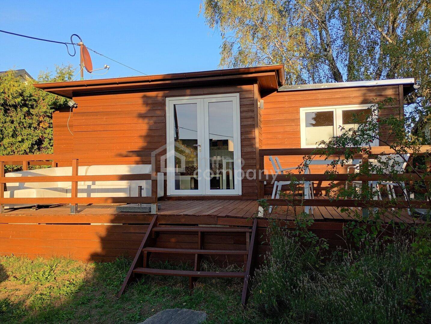 Kleines Holzhaus mit großer Holzterrasse und Zugang zum Gartenbereich, umgeben von Bäumen.