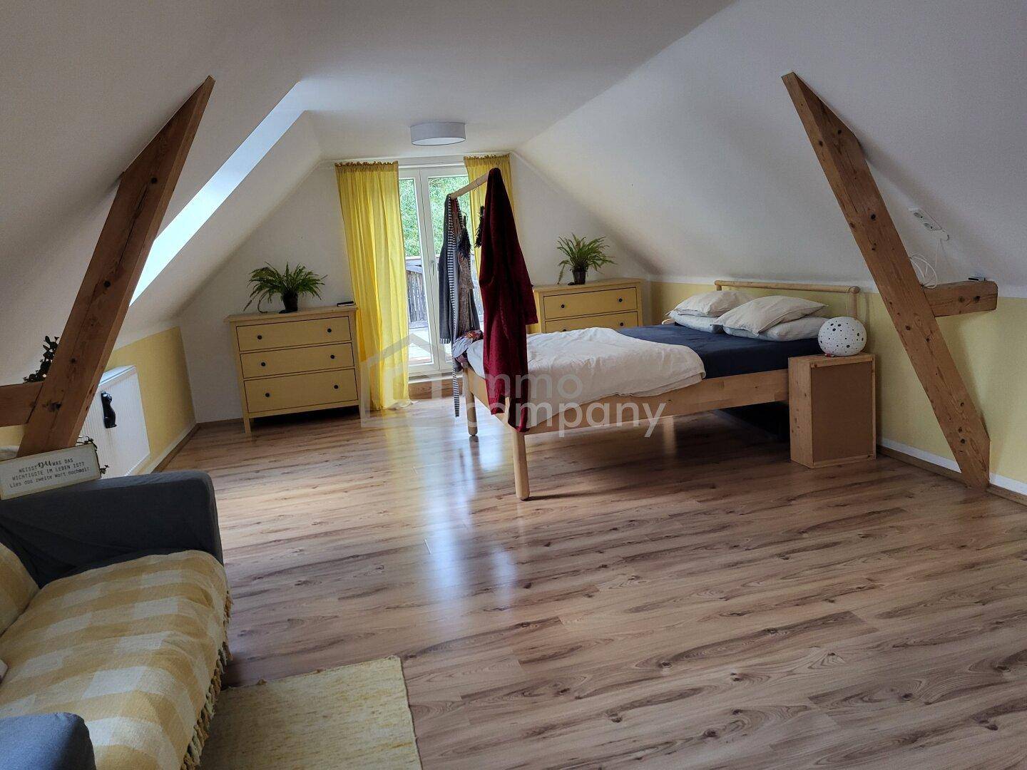 Helles Schlafzimmer im Dachgeschoss mit Holzbalken, Laminatboden und Zugang zum Balkon.