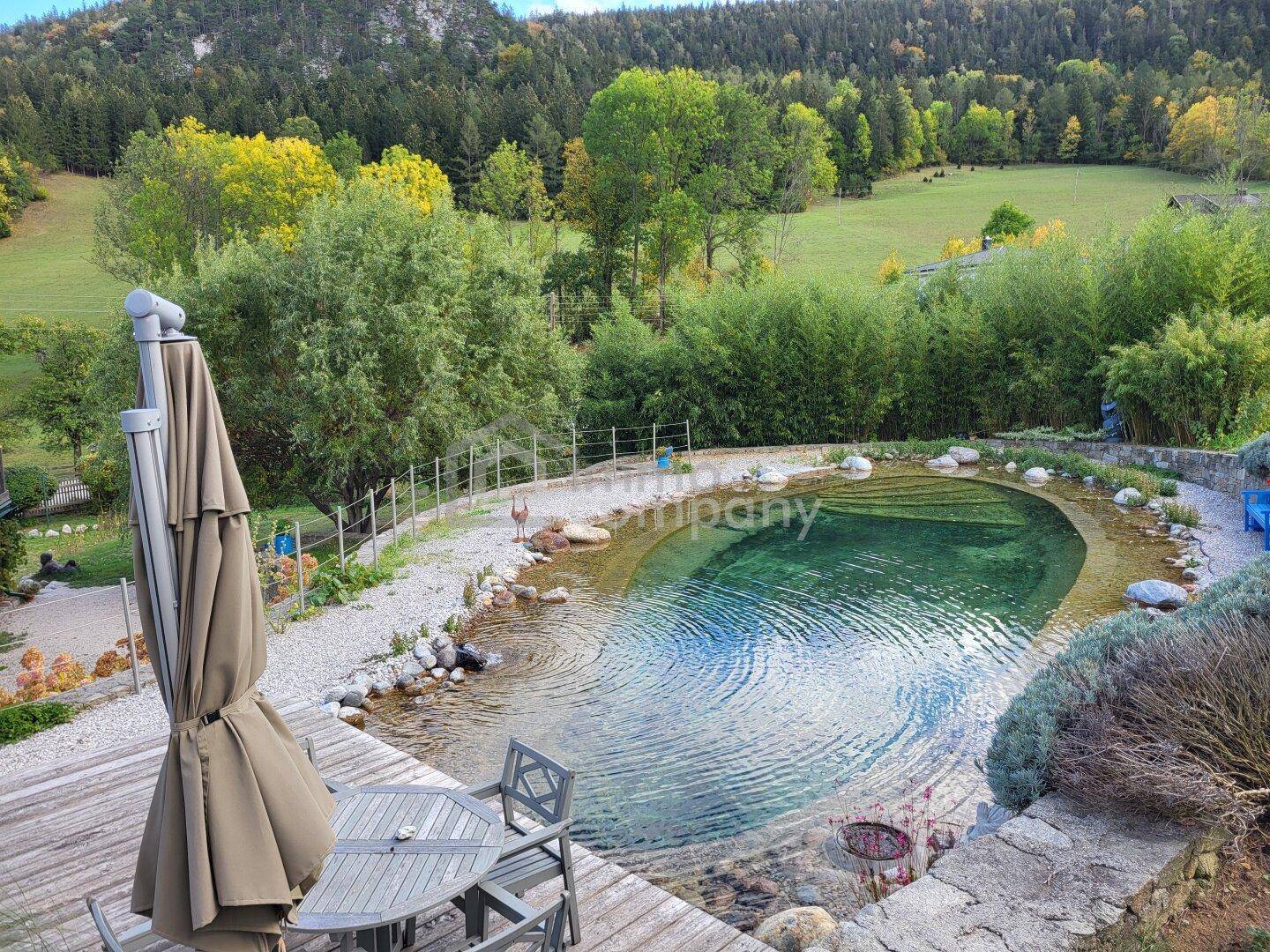 Holzterrasse mit Sitzgelegenheiten und einem natürlichen Schwimmteich, umgeben von grüner Landschaft.