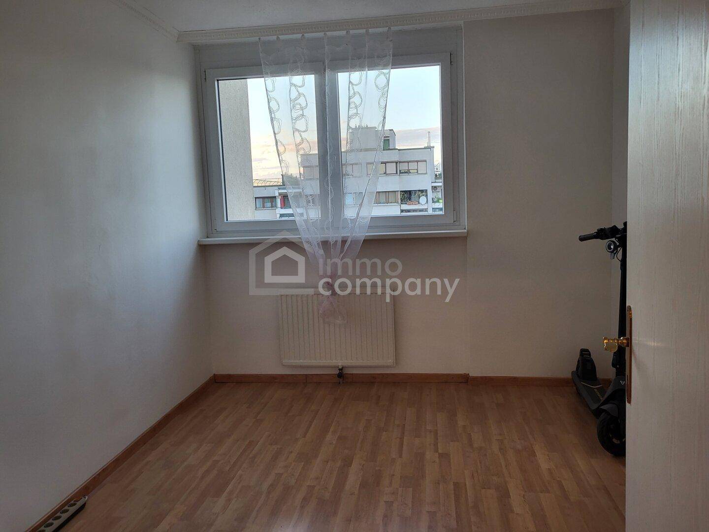 Leerer Raum mit hellem Laminatboden, weißen Wänden und einem Fenster mit Blick auf umliegende Gebäude.