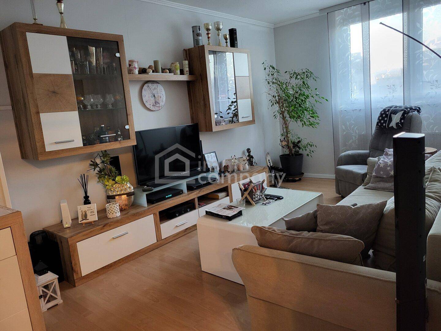 Helles Wohnzimmer mit beigem Sofa, modernem TV-Möbel und großen Fenstern mit Vorhängen.
