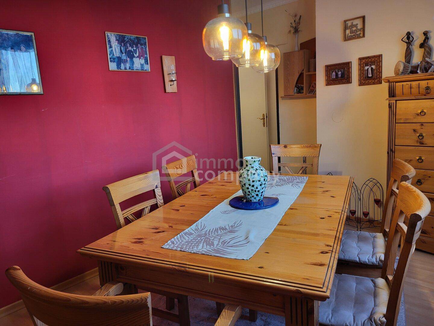 Gemütlicher Essbereich mit Holztisch, Stühlen und einer roten Akzentwand unter modernen Hängelampen.