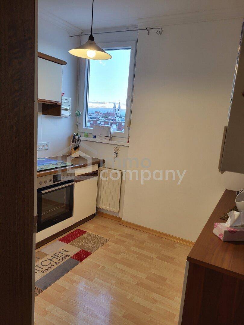 Kleine Küche mit hellem Laminatboden, weißen Schränken und Blick auf die Stadt durch das Fenster.
