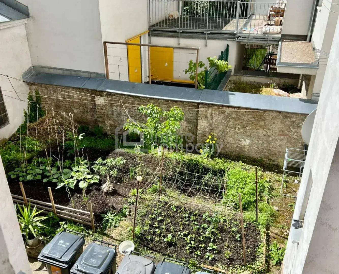 Kleiner, bepflanzter Innenhofgarten mit Hochbeeten und Mülltonnen, umgeben von Gebäuden.