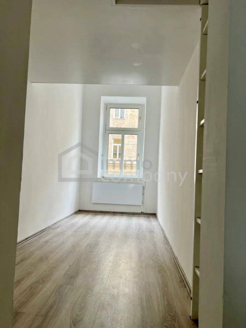 Heller, leerer Raum mit Laminatboden und einem Fenster, das auf ein Gebäude blickt.