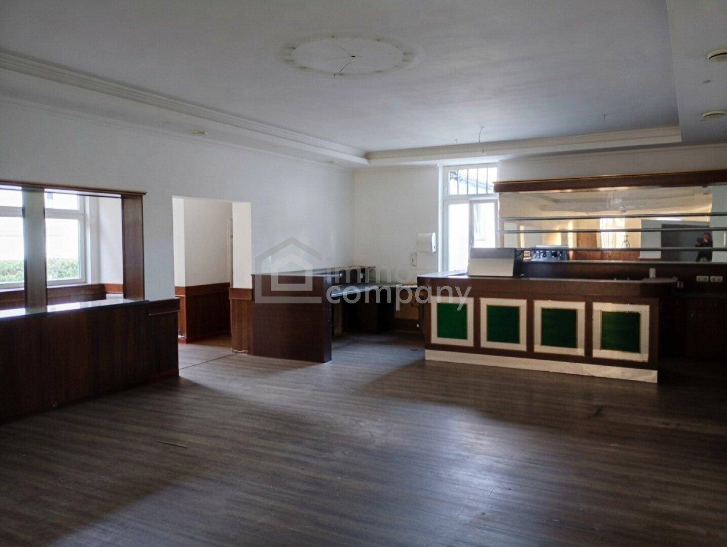 Geräumiger, leerer Raum mit Holzboden, einer Bar und großen Fenstern, ideal für Veranstaltungen.