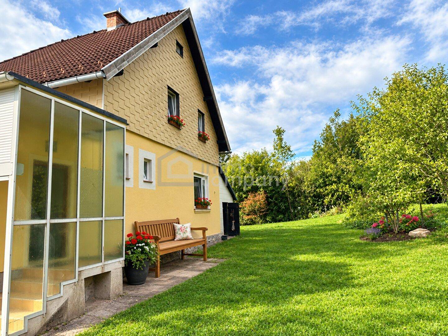 Gelbes Haus mit gepflegtem Garten, Wintergarten und Sitzbank unter blauem Himmel.