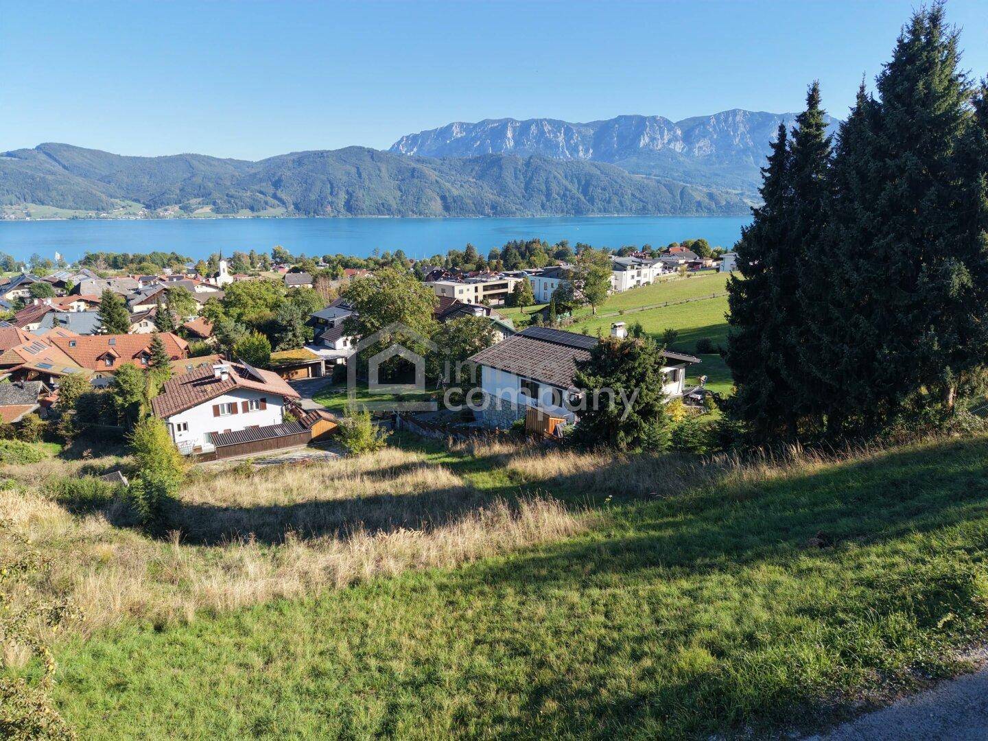 Weitläufiger Blick über eine Siedlung am See mit Bergen unter klarem blauem Himmel.