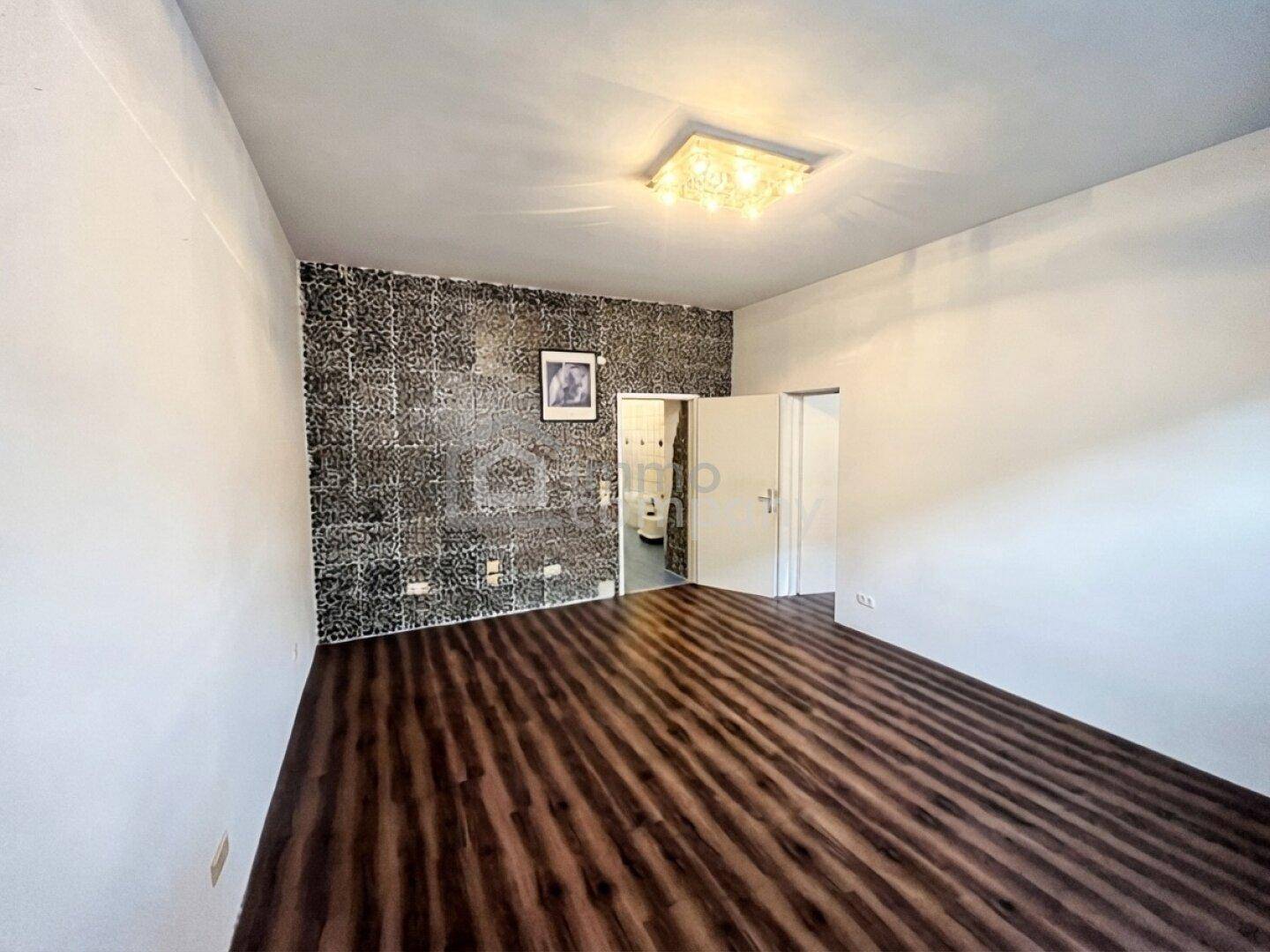 Leerer Raum mit dunklem Laminatboden, einer gemusterten Akzentwand und Zugang zu einem Badezimmer.