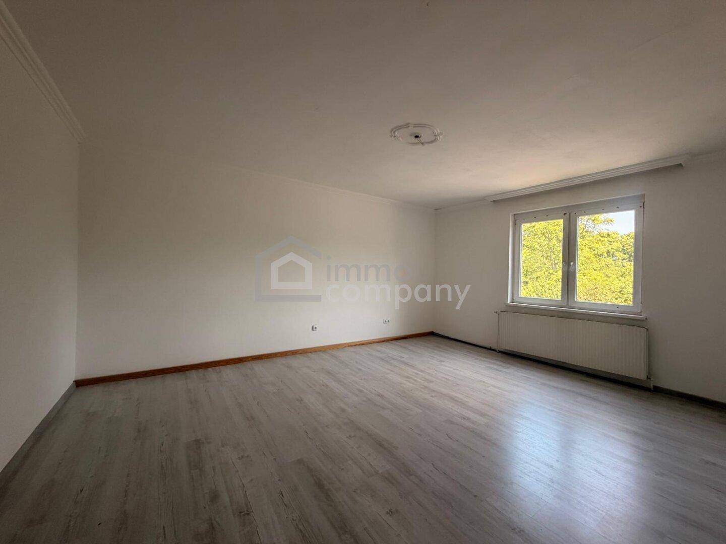 Leerer Raum mit hellen Wänden, Laminatboden und einem Fenster mit Heizkörper.