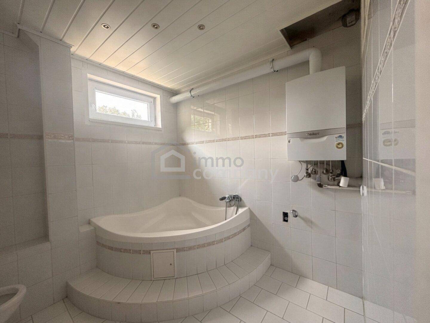 Badezimmer mit weißen Fliesen, Eckbadewanne mit Stufen und einem Fenster.