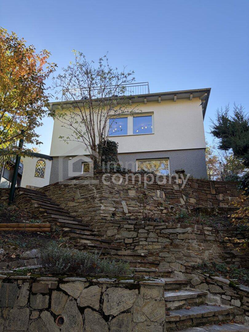 Rückseite des Hauses mit terrassierten Steinmauern und Treppen, umgeben von Bäumen.