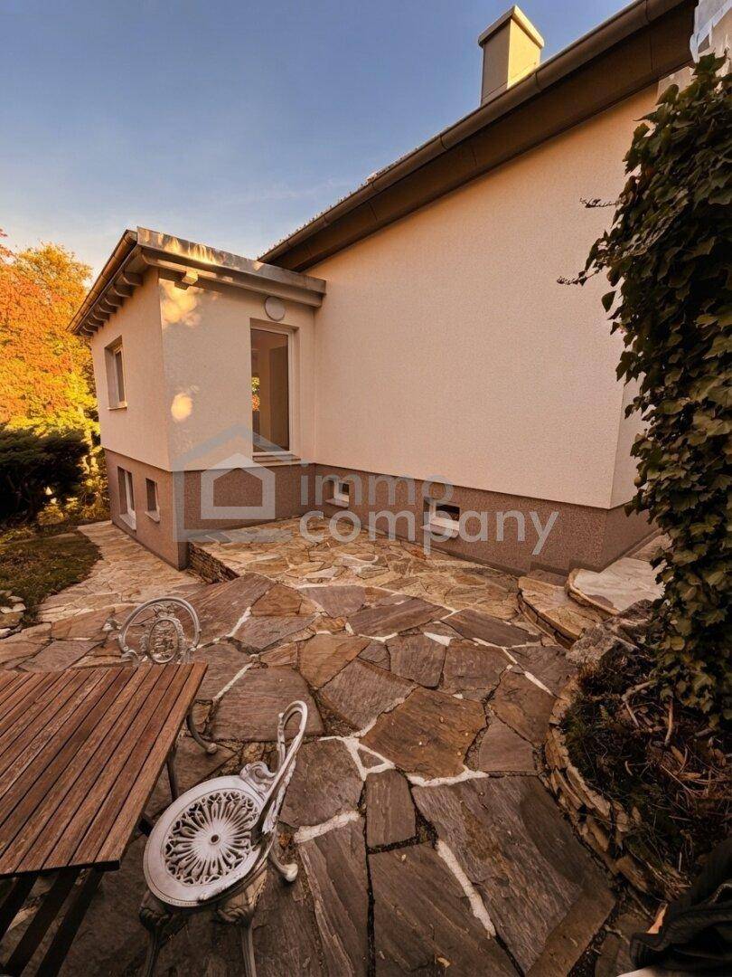 Seitlicher Blick auf das Haus mit großer Steinterrasse und Gartenmöbeln im Freien.