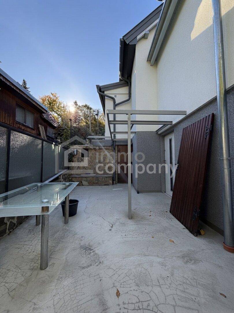 Gepflasterter Außenbereich neben dem Haus mit einem Glastisch und Überdachung.