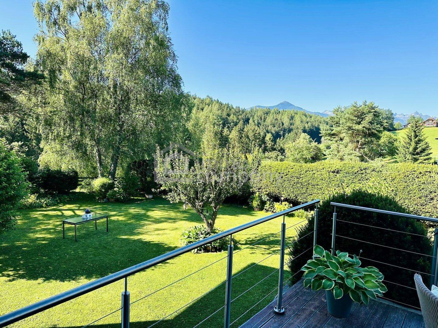 Sonniger Balkon mit Metallgeländer, Pflanzen und weitem Blick auf Berge und grüne Landschaft.
