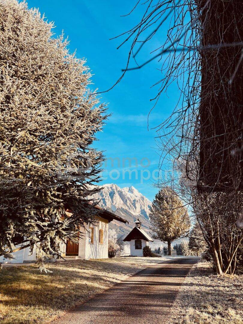 Winterliche Ansicht eines Anwesens mit verschneiten Bäumen, Kapelle und majestätischem Bergblick.