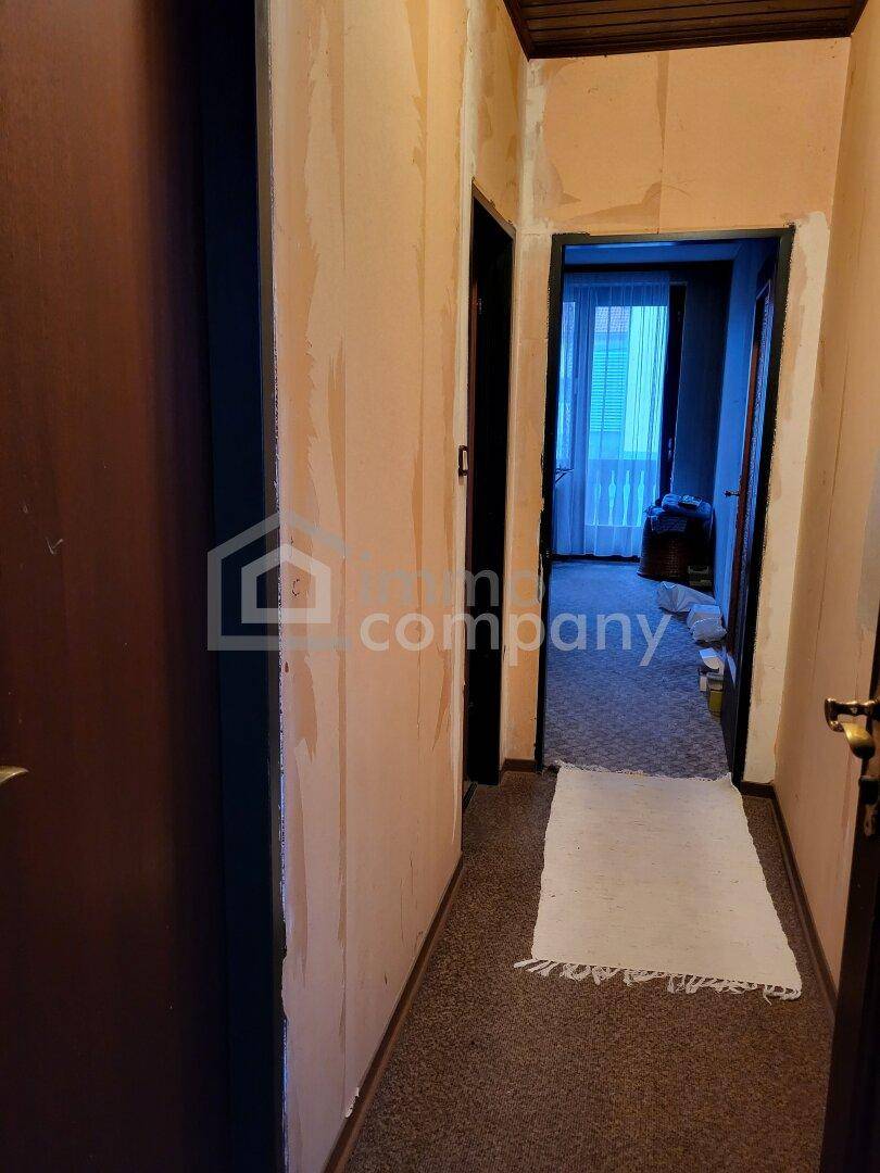 Schmaler Flur mit Teppichboden und teilweise abgelösten Tapeten, Blick in angrenzenden Raum.