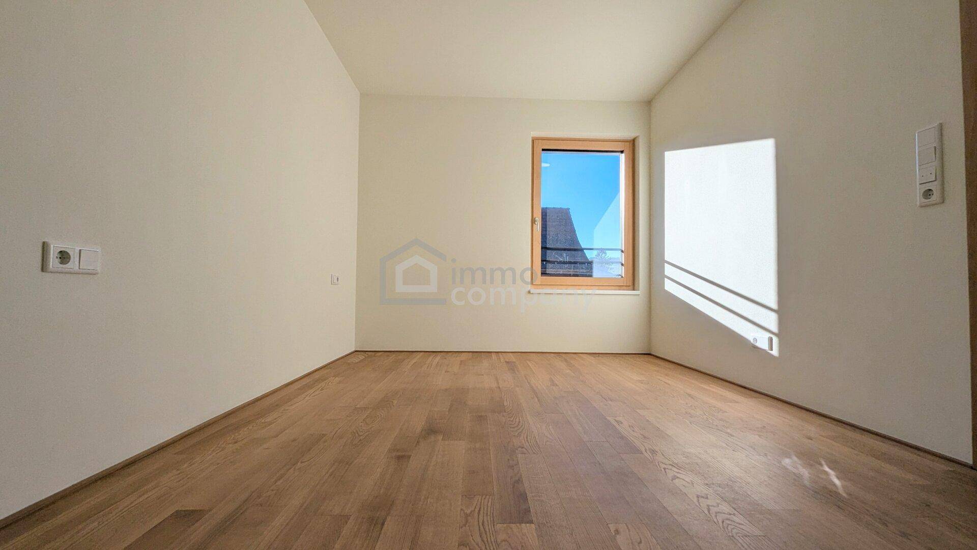 Leeres Schlafzimmer mit Holzboden und einem großen Fenster, das viel natürliches Licht hereinlässt.