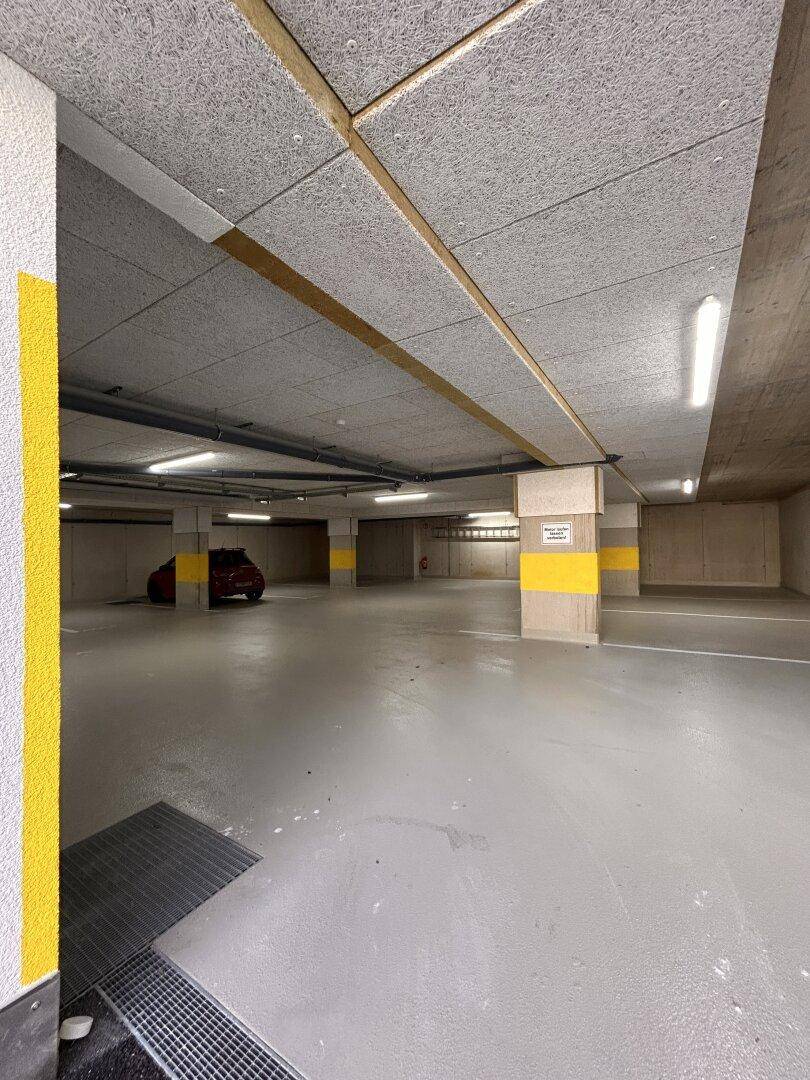 Helle Tiefgarage mit Betonstützen, gelben Markierungen und einem geparkten Fahrzeug im Hintergrund.