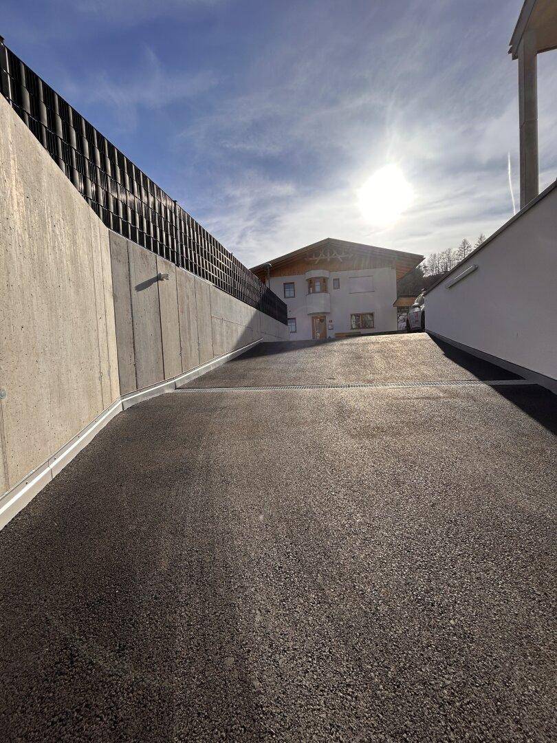 Asphaltierte Zufahrt zu einem Gebäude mit hohen Betonmauern unter strahlendem Sonnenschein.
