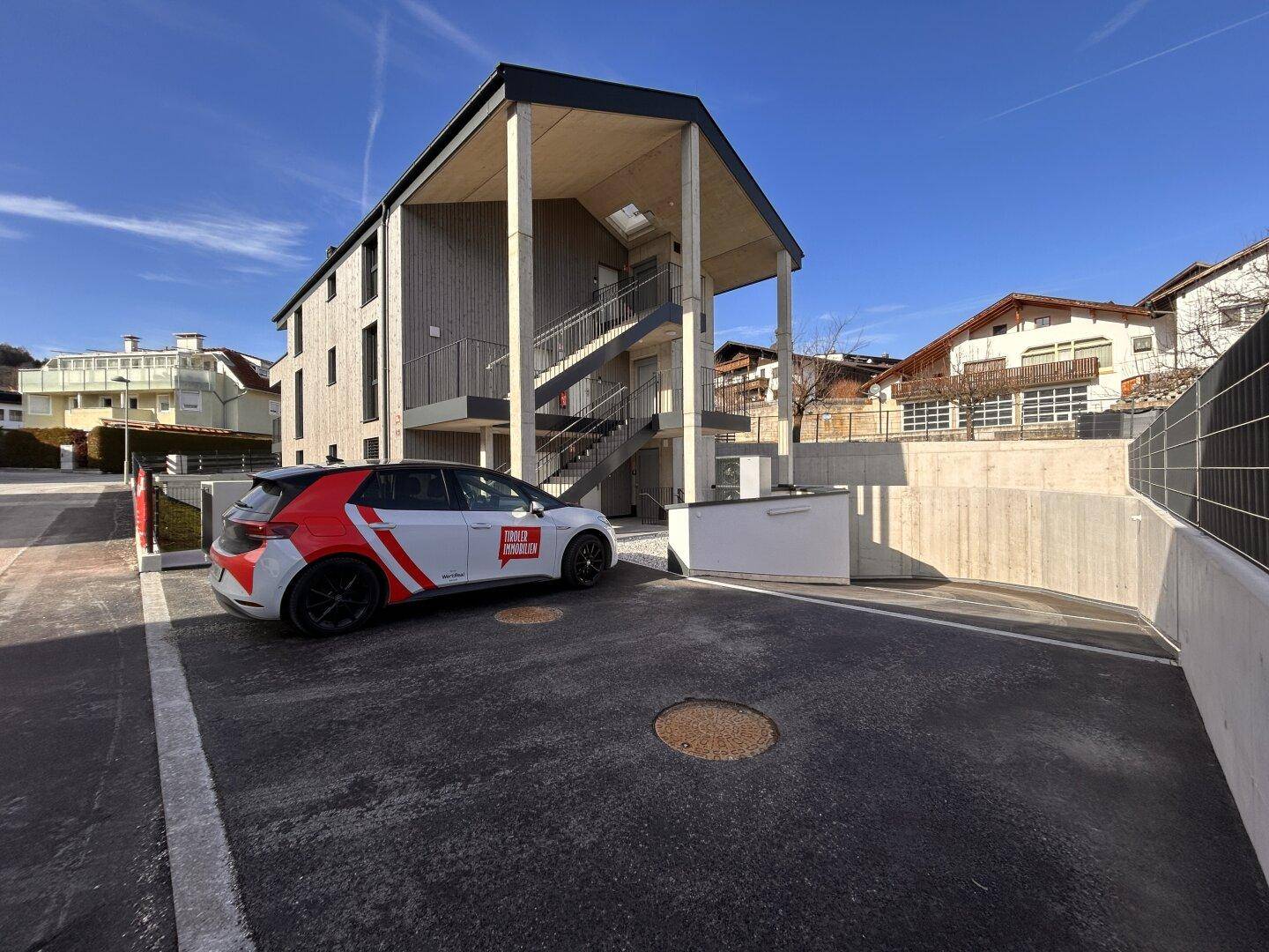 Modernes Gebäude mit Holzfassade und Außentreppe, davor ein geparktes Auto und die Tiefgaragenzufahrt.
